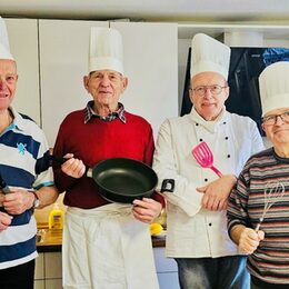 Das Bild zeigt vier ältere Herren. Alle mit einer Kochmütze auf dem Kopf und Küchengeräten in der Hand.